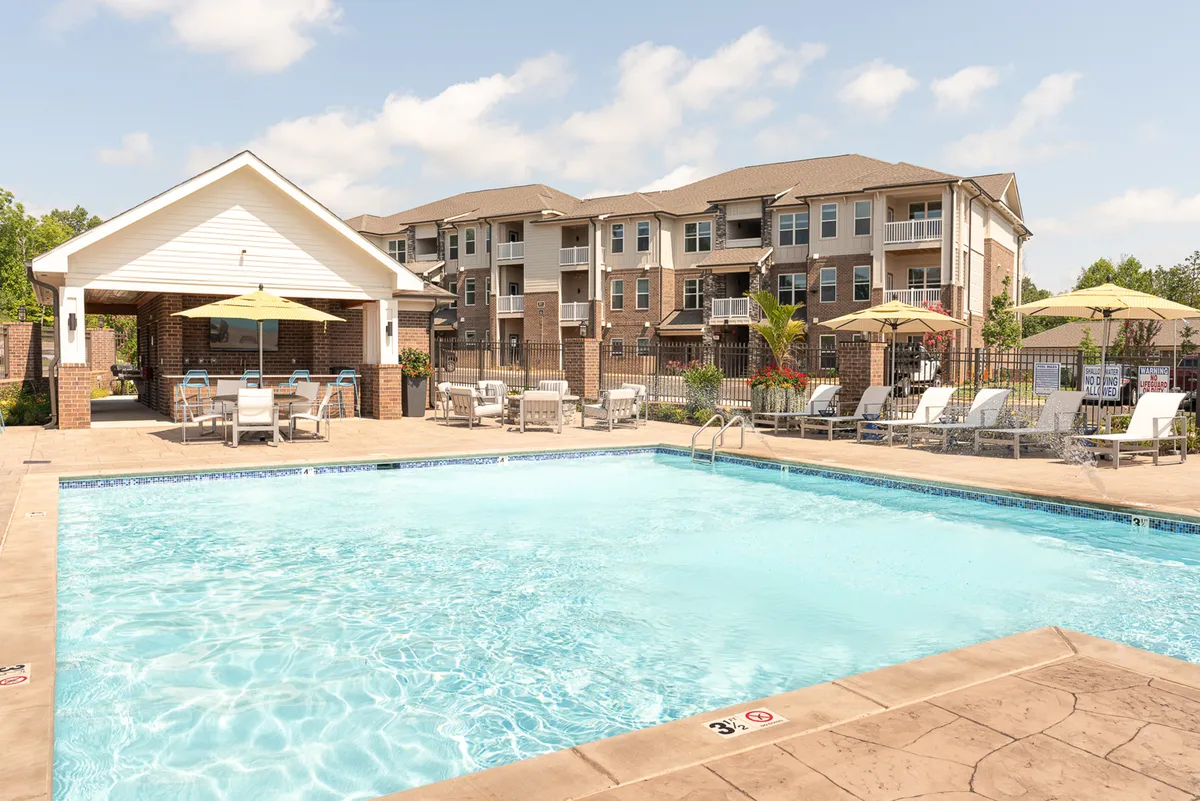 This exterior image showcases a sparkling community swimming pool surrounded by lounge chairs and umbrellas, with a covered seating area and the apartment buildings in the background. The scene is sunlit and inviting, suggesting both relaxation and social opportunities.