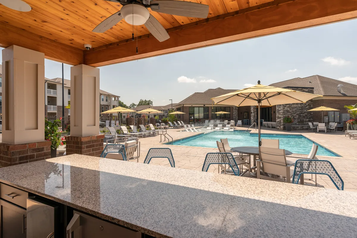 This vibrant exterior shot spotlights the sparkling swimming pool surrounded by comfortable outdoor seating, umbrellas, and a bar counter. The setup invites residents to socialize, unwind, or host casual gatherings in an inviting environment.