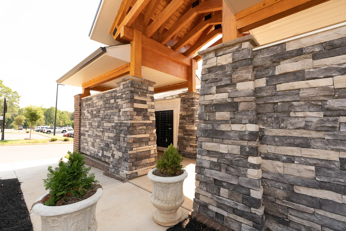 This exterior image features the welcoming entrance to the property, with rustic stone walls, wooden beams, and lush planters setting a warm and inviting tone. The outdoor area appears ready to greet both new arrivals and returning guests, hinting at a sense of community and comfort.