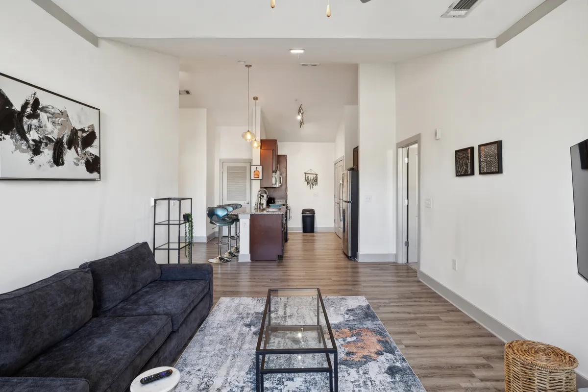 This image presents a wide view of an open-plan living and kitchen area. The scene features a modern sofa with a coffee table, stylish artwork adorning the wall, and a kitchen with contemporary finishes in the background. Natural light fills the space, highlighting the upscale furnishings and creating a welcoming atmosphere.