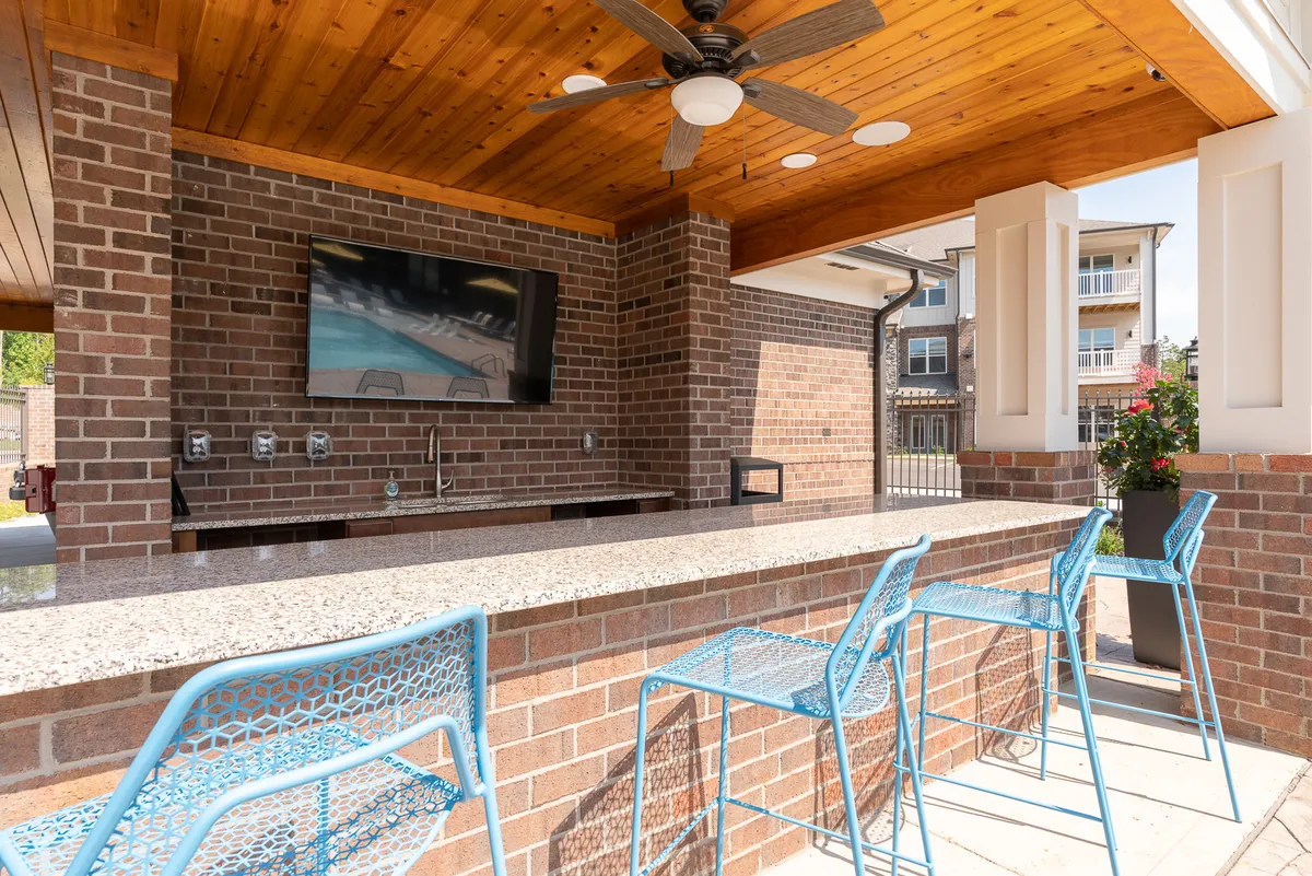 This exterior scene captures a lively communal space featuring a granite bar counter, blue metal chairs, a TV screen, and exposed brick walls. The area is set up for social gatherings and relaxation, suggesting opportunities for guests to mingle, enjoy meals outdoors, or catch a game together.