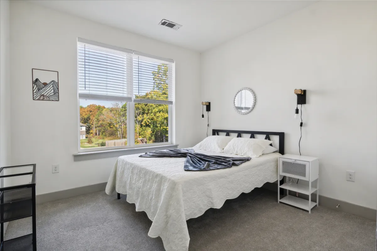 A sunlit bedroom with a cozy bed, a large window allowing natural light to stream in, a nightstand, and artistic decor. The scene feels inviting and serene, perfect for both restful sleep and quiet reading sessions.